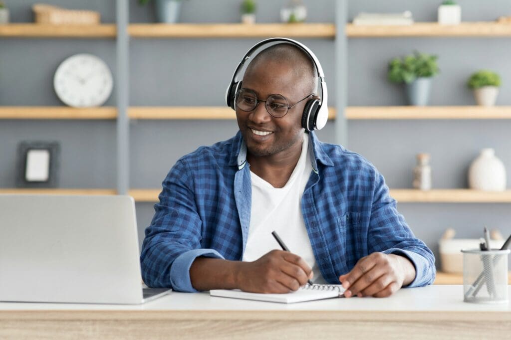 E-education. Happy black mature man using laptop and taking notes, listening online course